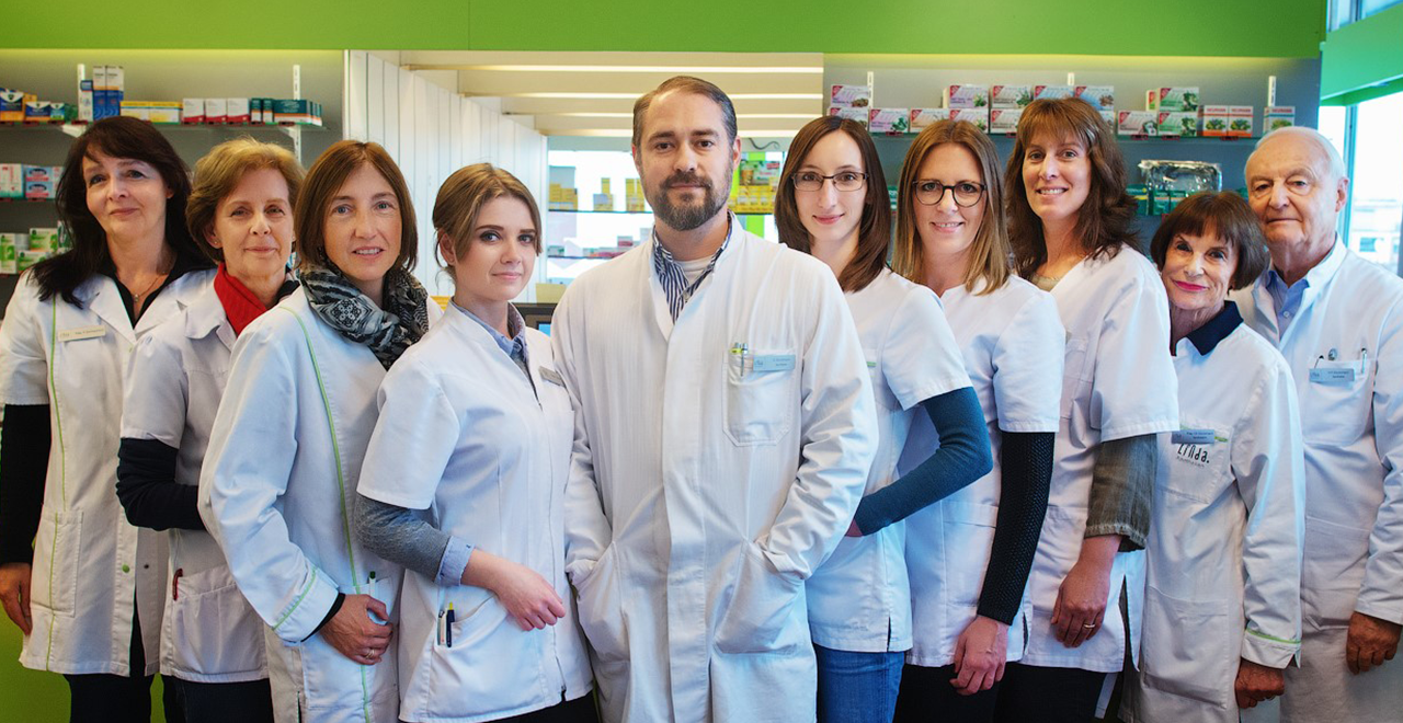 Gruppenfoto eines Apothekenteams mit zehn Mitarbeitenden in weißen Kitteln, die lächelnd in die Kamera blicken. Sie stehen in einer hellen, modernen Apothekenumgebung mit Regalen im Hintergrund.