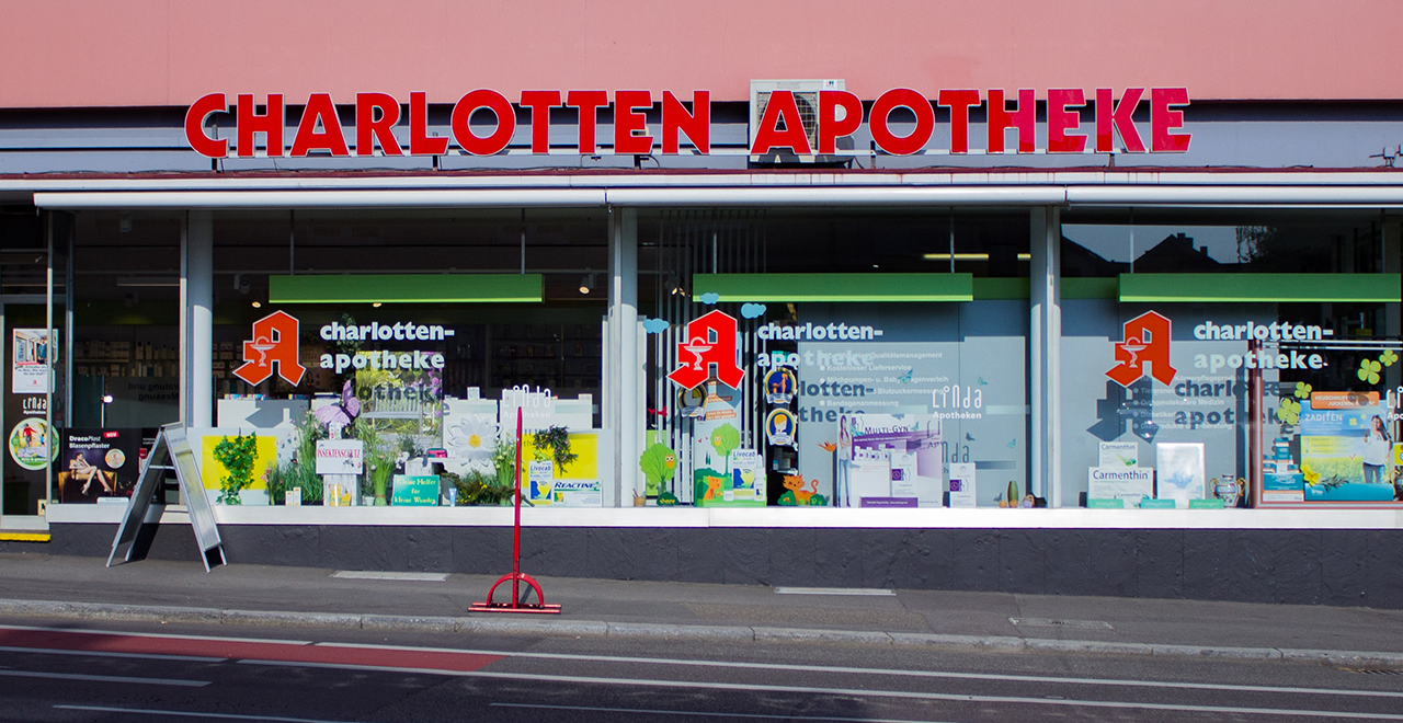 Fassade der Charlotten Apotheke mit großem roten Schriftzug über den Schaufenstern. Die Fenster sind mit Gesundheitsprodukten und bunten Dekorationen gestaltet. Der Eingangsbereich befindet sich links im Bild.