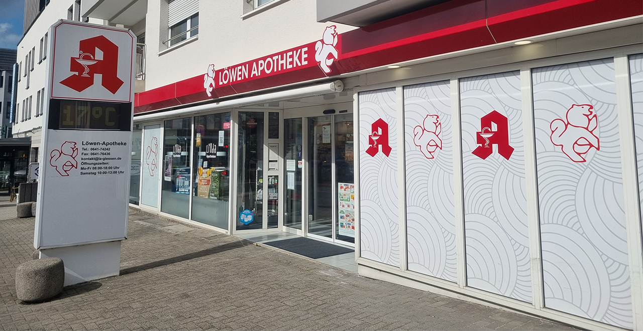 Außenansicht eines Apothekengebäudes mit einem auffälligen Schild, auf dem „Löwen Apotheke“ steht. Die Ladenfassade ist modern, mit einem weißen und roten Design, das ein Bild eines Löwen zeigt, der möglicherweise das Maskottchen der Apotheke darstellt. Große Fenster gewähren Einblick in die Apotheke, und es gibt weitere Schilder auf den Fenstern, die wahrscheinlich Dienstleistungen oder Sonderaktionen bewerben. Der Eingang ist deutlich markiert, und der Bürgersteig ist sauber und gepflegt.