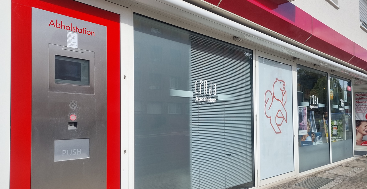 Außenansicht einer Apotheke mit einem Selbstabholstation auf der linken Seite des Eingangs. Die Maschine ist silbern mit roten Akzenten und mit „Abholstation“ beschriftet. Das Logo der Apotheke ist durch das Fenster sichtbar, und der Eingang ist mit Jalousien versehen. Es gibt auch ein großes Schild, das auf die Abholstation hinweist.