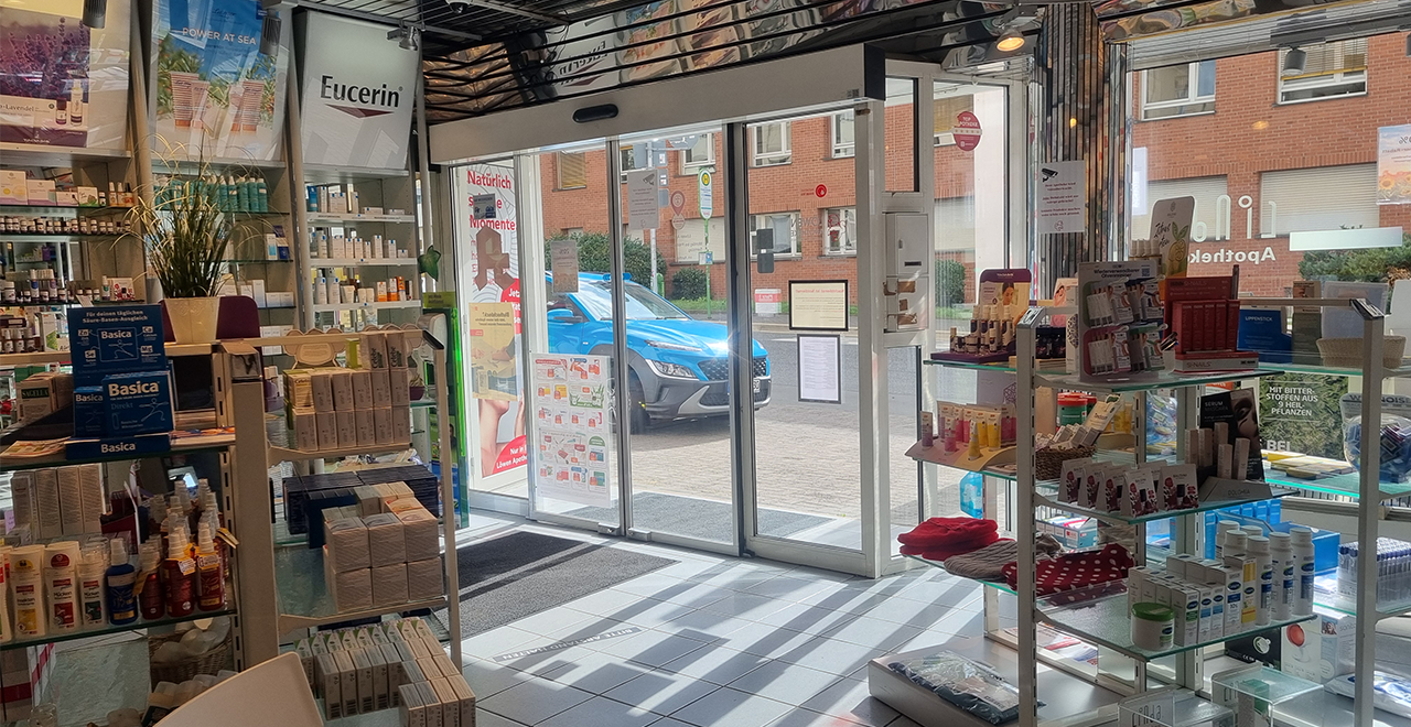 Ein Blick in die Apotheke, die von einem modernen, offenen Raum geprägt ist. Auf der linken Seite sind Regale mit Gesundheits- und Pflegeprodukten wie Lotionen, Medikamenten und Nahrungsergänzungsmitteln sichtbar. In der Mitte führt eine automatische Schiebetür nach draußen, wo ein blaues Auto vor der Apotheke steht. Der Eingangsbereich ist gut beleuchtet, und auf der rechten Seite sind weitere Regale mit ähnlichen Produkten. An den Wänden hängen Werbeplakate für natürliche Produkte.