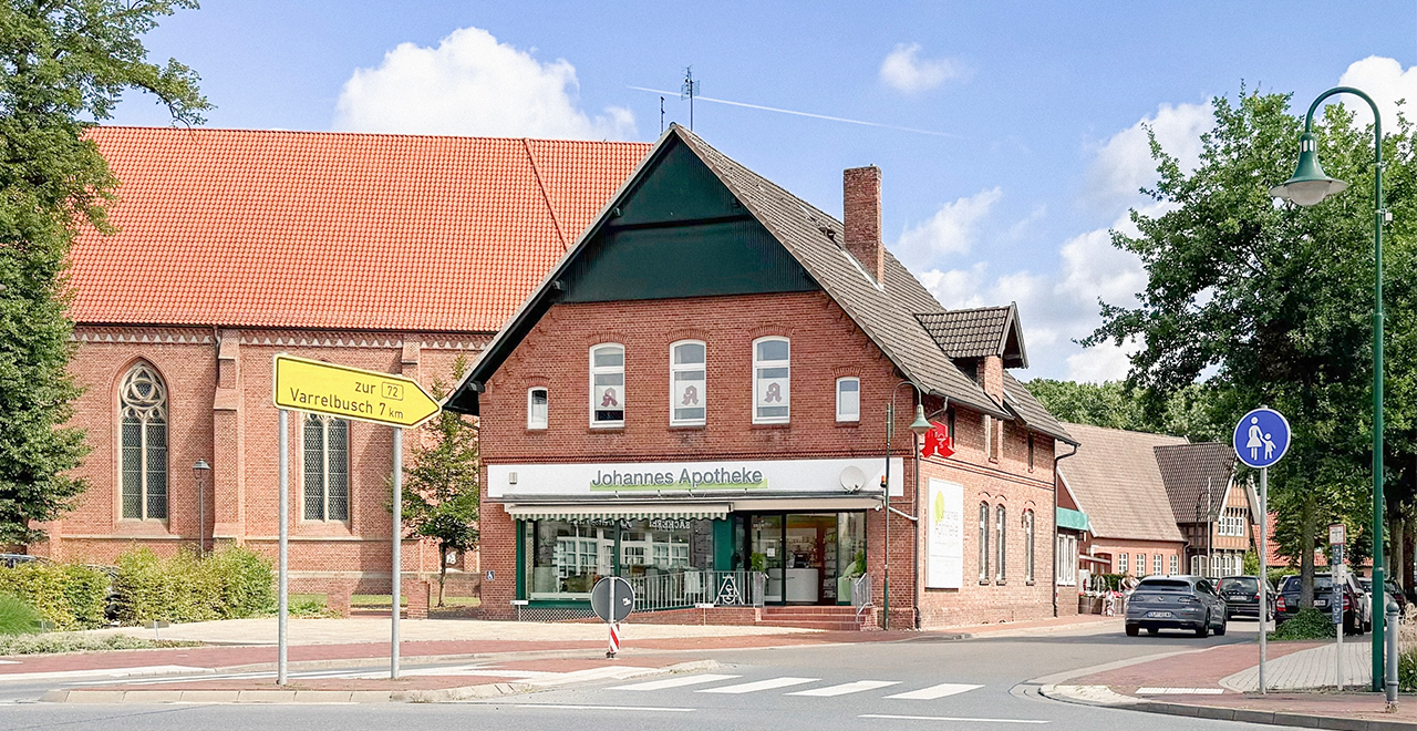 Außenansicht der Johannes Apotheke in einem roten Backsteingebäude an einer Straßenecke mit barrierefreiem Eingang und angrenzender Kirche im Hintergrund.