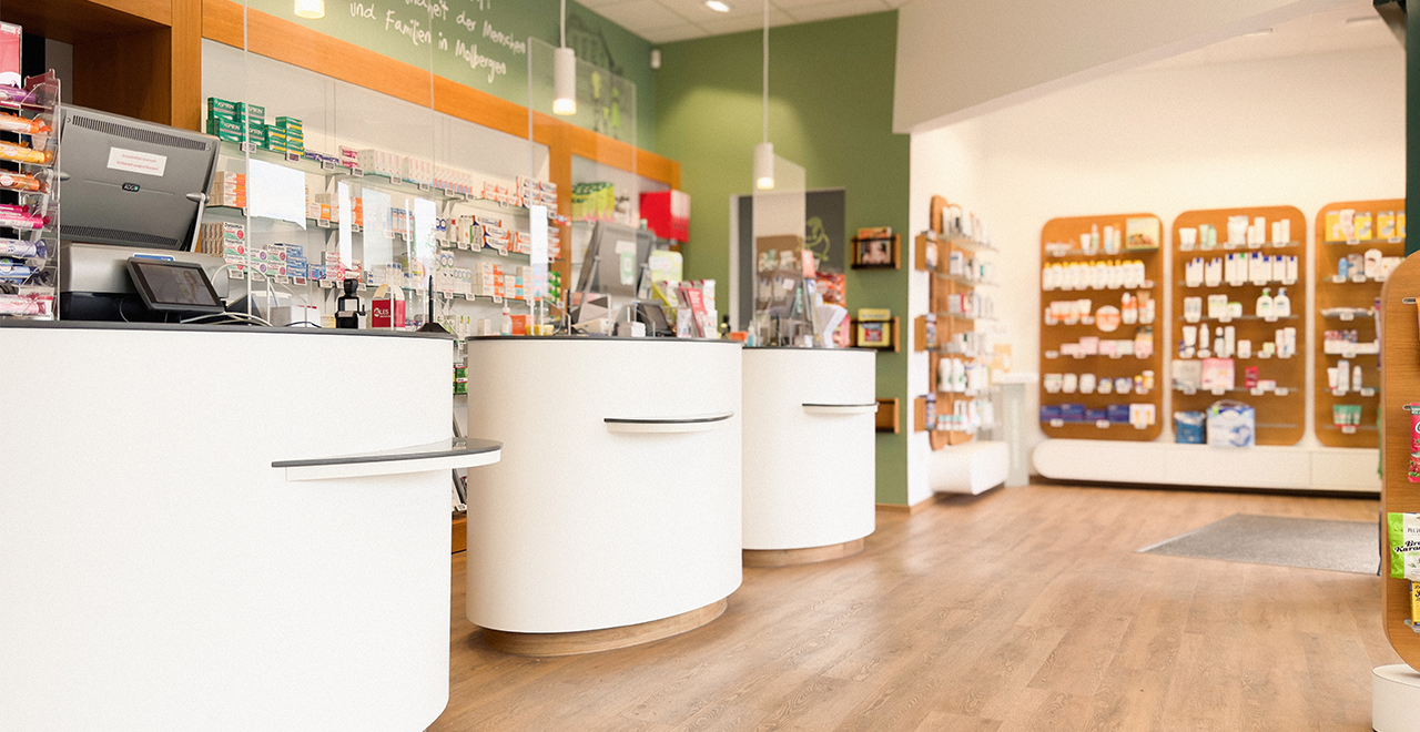 Übersichtlicher Innenraum einer Apotheke mit weißen Theken, hölzernen Wandregalen voller Produkte und einem modernen, offenen Raumkonzept.