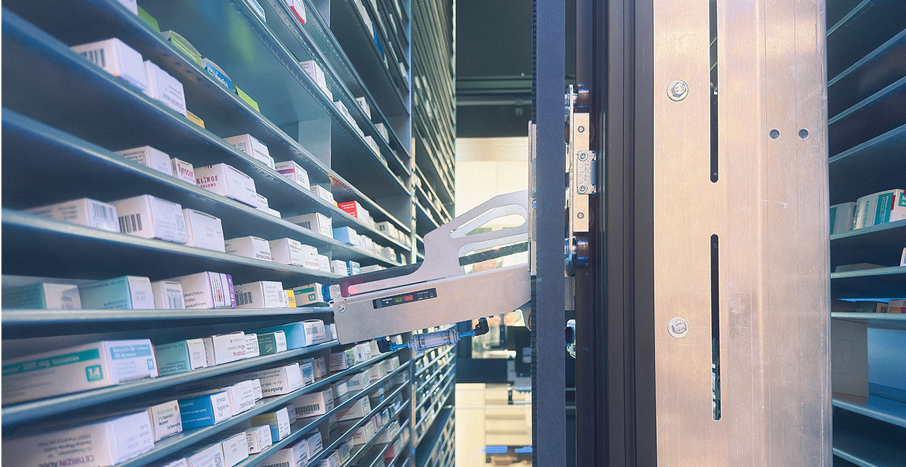 Automatisiertes Apothekenlager mit Kommissionierroboter, der Medikamentenschachteln aus hohen, schmalen Regalen entnimmt.
