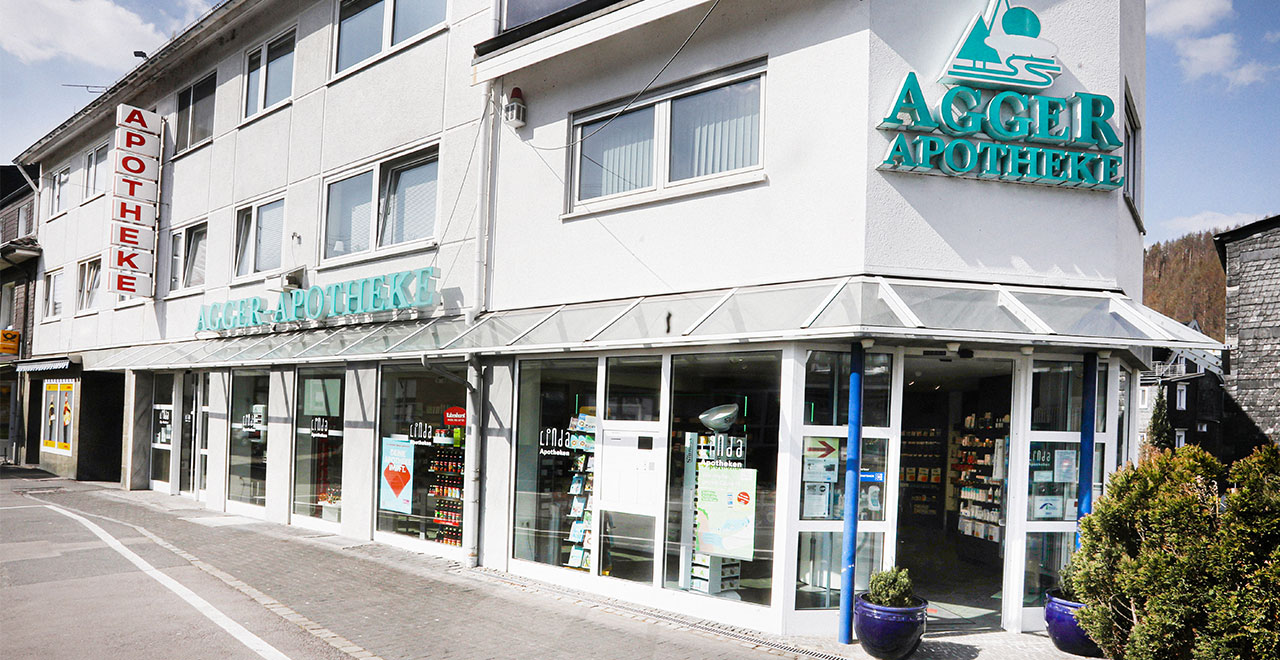 Außenansicht der Agger-Apotheke in einem mehrstöckigen Gebäude.
Über den großflächigen Schaufenstern sind der Schriftzug „Agger-Apotheke“ und weitere Apothekenschilder angebracht. Der Eingang befindet sich auf der Straßenseite.