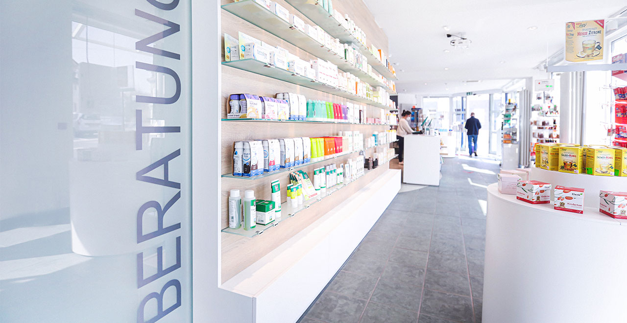 Innenansicht der Agger-Apotheke mit langen Regalen, in denen Arzneimittel, Kosmetik- und Pflegeprodukte übersichtlich präsentiert werden. Im Hintergrund befindet sich der Verkaufstresen.