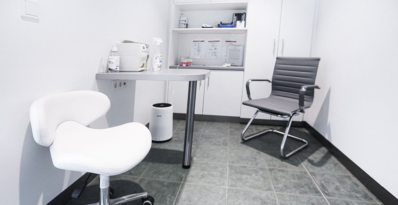 Separater Beratungsraum in einer Apotheke mit weißem Untersuchungsstuhl, Schreibtisch, schwarzem Besucherstuhl und weißen Einbauschränken.
Der Raum ist hell gestaltet und mit grauen Bodenfliesen ausgestattet.