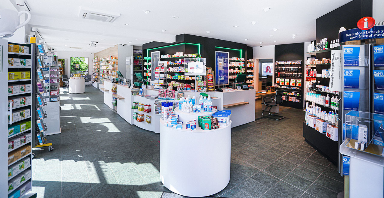 Weitläufiger Blick in eine moderne Apotheke mit weißen Verkaufstresen,
Produktregalen und einer runden Präsentationsfläche im Vordergrund.
Im Hintergrund befinden sich weitere Regale mit grüner Beleuchtung sowie ein Beratungsbereich.
Tageslicht fällt durch die Fenster im Eingangsbereich.