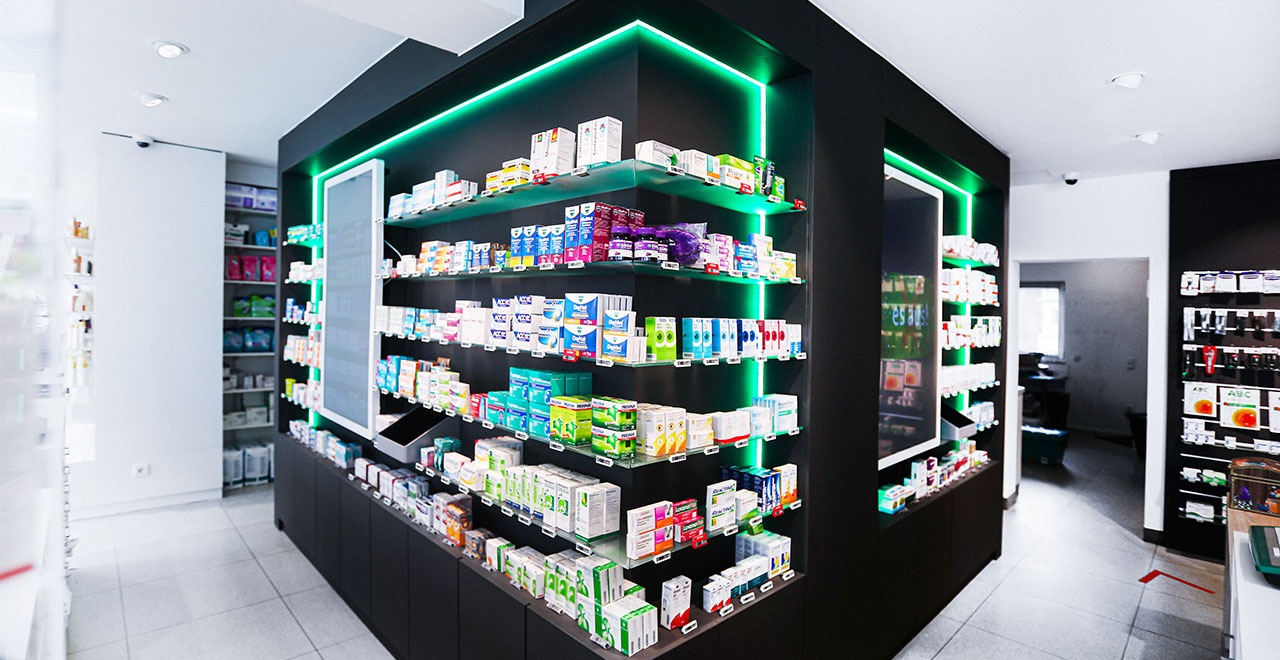 Nahaufnahme eines schwarzen Regalsystems in einer Apotheke mit mehreren Fächern voller Arzneimittelpackungen. Grüne LED-Lichtleisten umranden die Regale. Im Vordergrund ist der geflieste Verkaufsraum zu sehen.