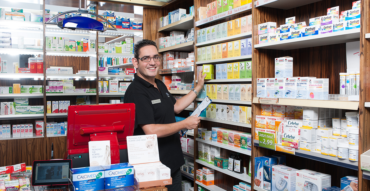 Ein Apotheker steht an einem Regal in einer Apotheke und sortiert Produkte. Das Regal ist voll mit Medikamenten und Nahrungsergänzungsmitteln. Der Apotheker trägt ein schwarzes Polo-Shirt und lächelt in die Kamera. Im Vordergrund ist die Kasse mit einem roten Bildschirm und einem Laptop zu sehen. Auf dem Tisch vor ihm befinden sich einige Produkte und Flyer.