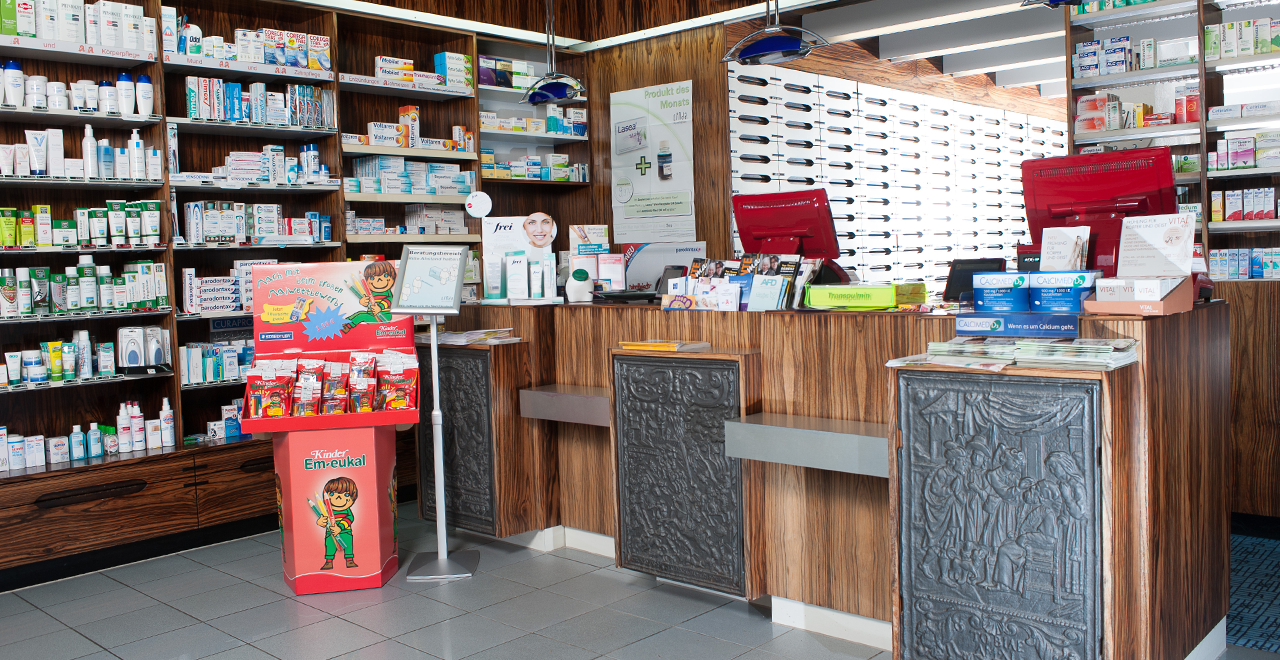 Innenansicht einer modernen Apotheke. Der Verkaufstresen ist aus Holz mit eingelassenen Regalen für Produkte. An den Wänden sind Regale mit verschiedenen Gesundheitsprodukten wie Hautpflege und Medikamenten zu sehen. Ein Verkaufsständer mit bunten Verpackungen von „Kinder Em-eukal“ steht im Vordergrund. An den Wänden hängen Informationsblätter und Werbung für Produkte des Monats. Es gibt auch digitale Bildschirme und Kassenbereiche im Hintergrund.