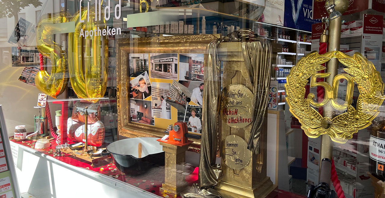 Schaufenster der Kreuz Apotheke zum 50-jährigen Jubiläum, dekoriert mit goldenen Luftballons in Form der Zahl 50, goldfarbenen Ausstellungsobjekten und einer Bildercollage zur Geschichte der Apotheke.