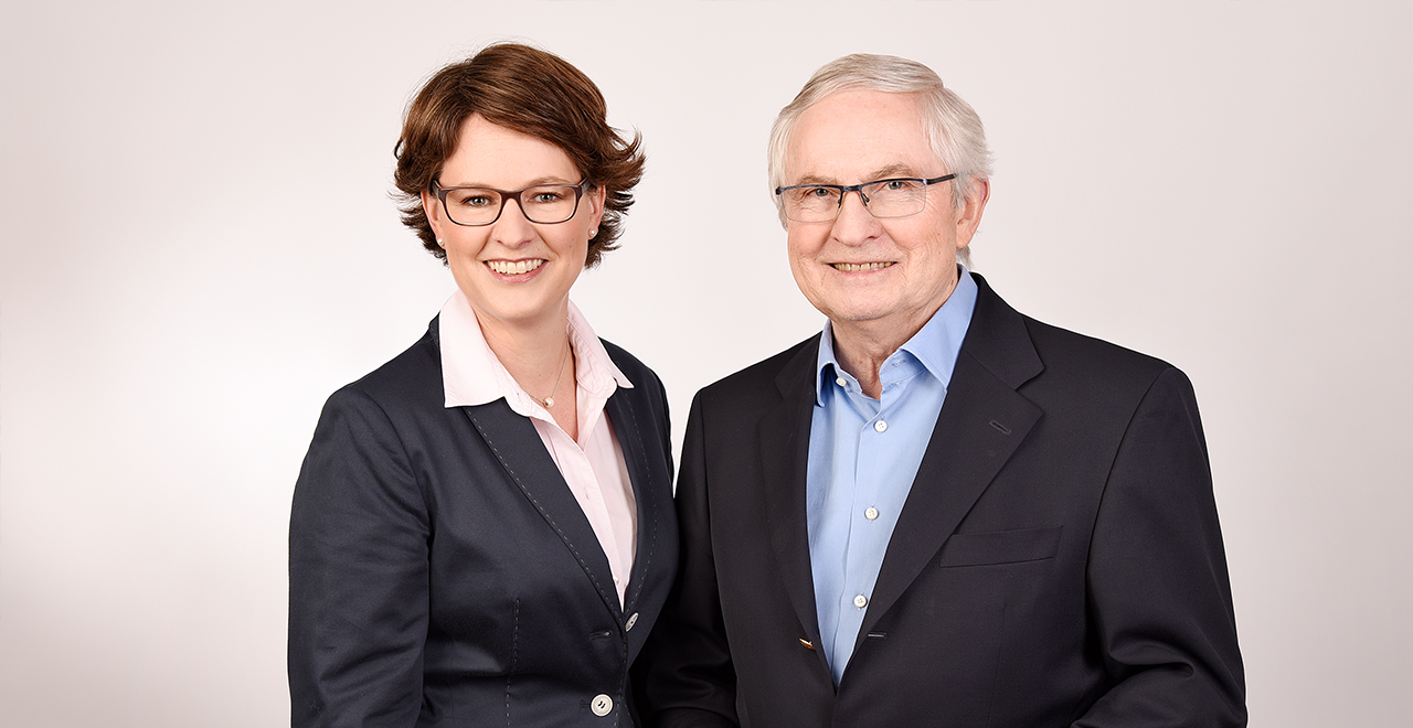 Porträtfoto des Inhabers der Kreuz-Apotheke und seiner Tochter vor hellem Hintergrund. Beide tragen Brillen und dunkle Blazer. Die Tochter hat kurze braune Haare, ein weißes Hemd mit rosa Kragen und lächelt freundlich. Der Vater hat graue Haare, ein hellblaues Hemd unter einem dunklen Sakko und lächelt ebenfalls. Die Tochter unterstützt ihren Vater in der Leitung der Apotheke.