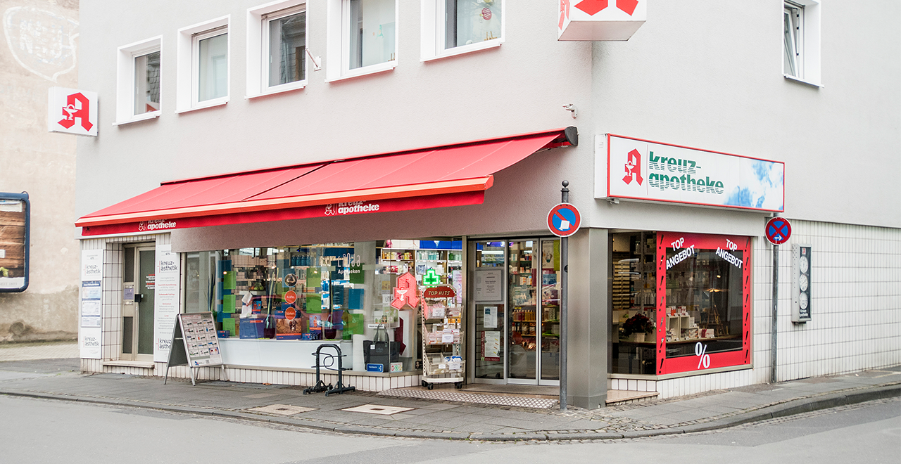 Außenansicht einer deutschen Apotheke an einer Straßenecke. Die Apotheke heißt „Kreuz-Apotheke“ und hat ein rotes Apothekensymbol über dem Eingang. Eine rote Markise ragt über die große Fensterfront, in der verschiedene Produkte ausgestellt sind. Rechts neben dem Haupteingang befindet sich ein weiteres Schaufenster mit der Aufschrift „Top Angebot“. Zwei Verkehrsschilder mit Halteverbot sind am Straßenrand zu sehen. Das Gebäude ist hell verputzt mit mehreren Fenstern im oberen Stockwerk.