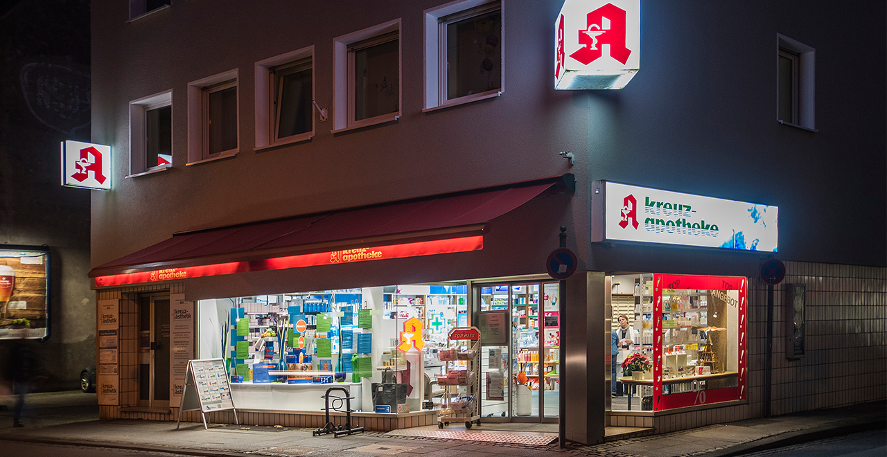 Nachtaufnahme einer deutschen Apotheke an einer Straßenecke. Die Apotheke heißt „Kreuz-Apotheke“ und ist hell beleuchtet. Leuchtschilder mit dem roten Apotheken-A und dem Namen der Apotheke sind deutlich sichtbar. Eine rote Markise erstreckt sich über das Schaufenster, das mit verschiedenen Produkten dekoriert ist. Im rechten Schaufenster steht „Top Angebot“ mit roten Prozentzeichen. Die Apotheke wirkt einladend und offen, auch eine Person ist im Inneren zu sehen. Die Umgebung ist dunkel, nur die Apotheke selbst ist gut ausgeleuchtet.