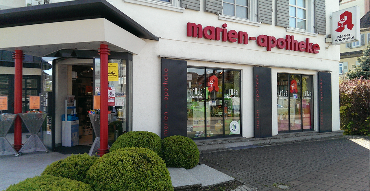 Außenansicht der Marien-Apotheke. Der Eingang ist von zwei roten Säulen eingerahmt, die das moderne Design des Gebäudes betonen. Über dem Eingang befindet sich der Schriftzug „marien-apotheke“ in roten Buchstaben. Durch die großen Fenster im Schaufenster sind verschiedene Produkte und Regale sichtbar. Auf der linken Seite befinden sich Werbeplakate, die Informationen zu Angeboten bieten.