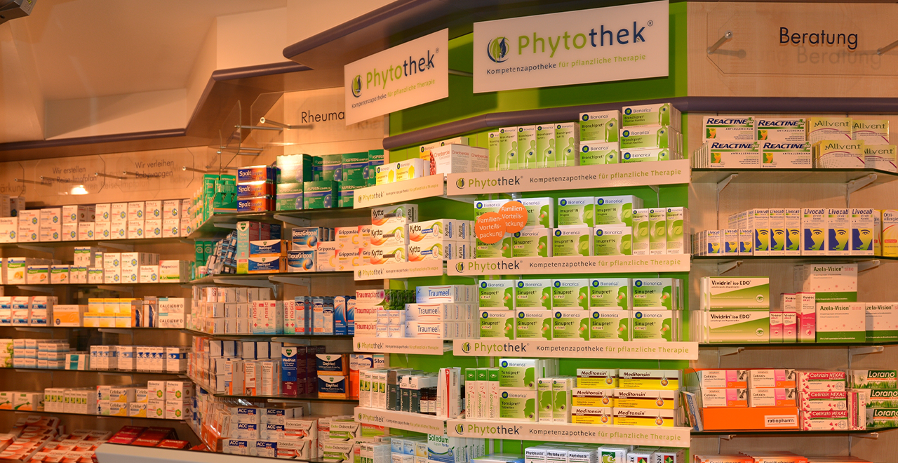 Ein Regal in der Apotheke mit einer Vielzahl von Produkten für die pflanzliche Therapie. In der Mitte des Regals steht der Schriftzug „Phytohek“, der auf die Spezialisierung der Apotheke für pflanzliche Arzneimittel hinweist. Die Regale sind voll mit verschiedenen Medikamenten und Nahrungsergänzungsmitteln, darunter auch Produkte der Marken Bionorica, Reactine und Livocab. An der Wand hinter dem Regal befinden sich Hinweise zu Beratungsmöglichkeiten und Rheuma-Spezialbehandlungen.
