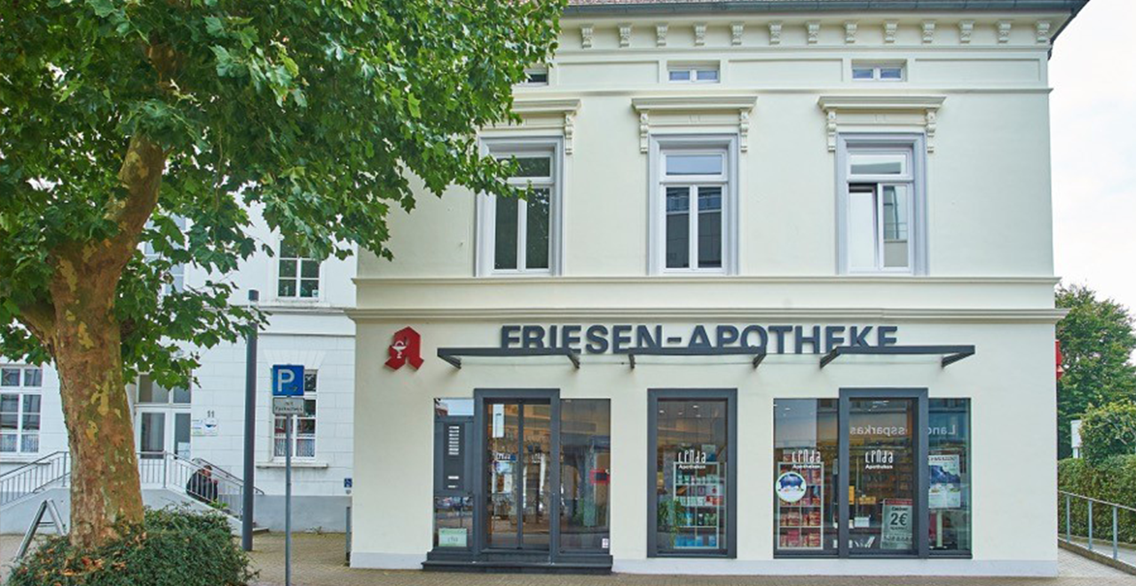 Außenansicht der Friesen-Apotheke. Das Gebäude hat eine helle Fassade und befindet sich in einer städtischen Umgebung mit Bäumen und Parkplätzen. Über dem Eingang ist der Schriftzug „FRIESEN-APOTHEKE“ in schwarzen Buchstaben zu sehen. Das Schaufenster zeigt verschiedene Produkte, und das Apotheken-Logo ist in rotem Farbton an der Ecke des Gebäudes angebracht.