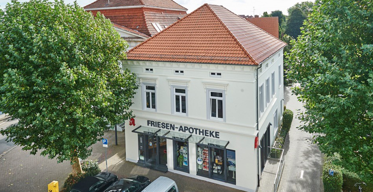 Blick auf das Gebäude der Friesen-Apotheke aus einer höheren Perspektive. Der Schriftzug „FRIESEN-APOTHEKE“ ist gut sichtbar, ebenso wie das rote Apotheken-Logo. Um das Gebäude herum sind mehrere Bäume und parkende Autos zu sehen, während das Gebäude im Hintergrund eine klassische Architektur aufweist.