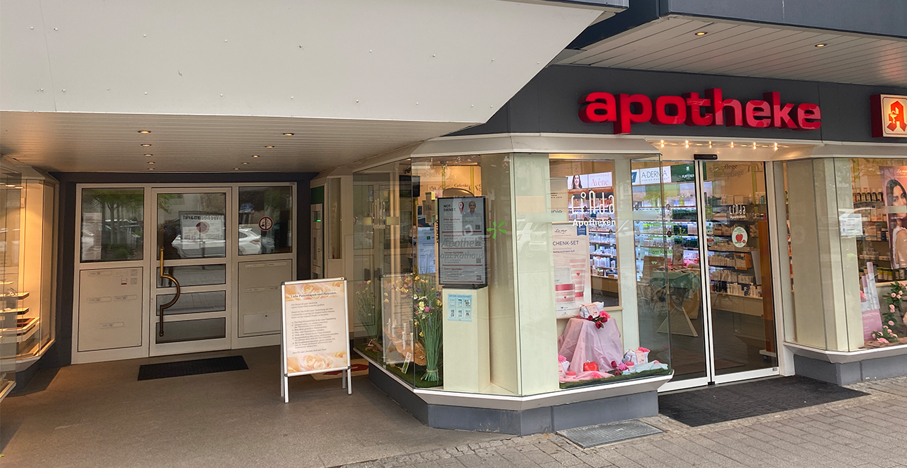 Eingangsbereich einer Apotheke im Erdgeschoss eines Ärztehauses mit dem Schriftzug „Apotheke“ in roten Buchstaben. Über dem Eingang ist ein großes Schild mit der Adresse „Rathausplatz 4“ und Hinweis auf ein Ärztehaus.
