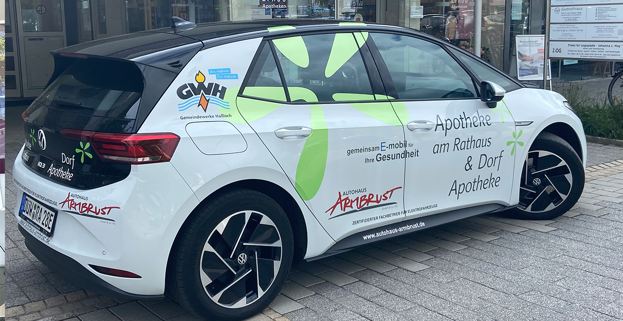 Ein weißes Elektroauto mit Werbeaufdruck „Apotheke am Rathaus & Dorf Apotheke“ steht vor der Apotheke. 