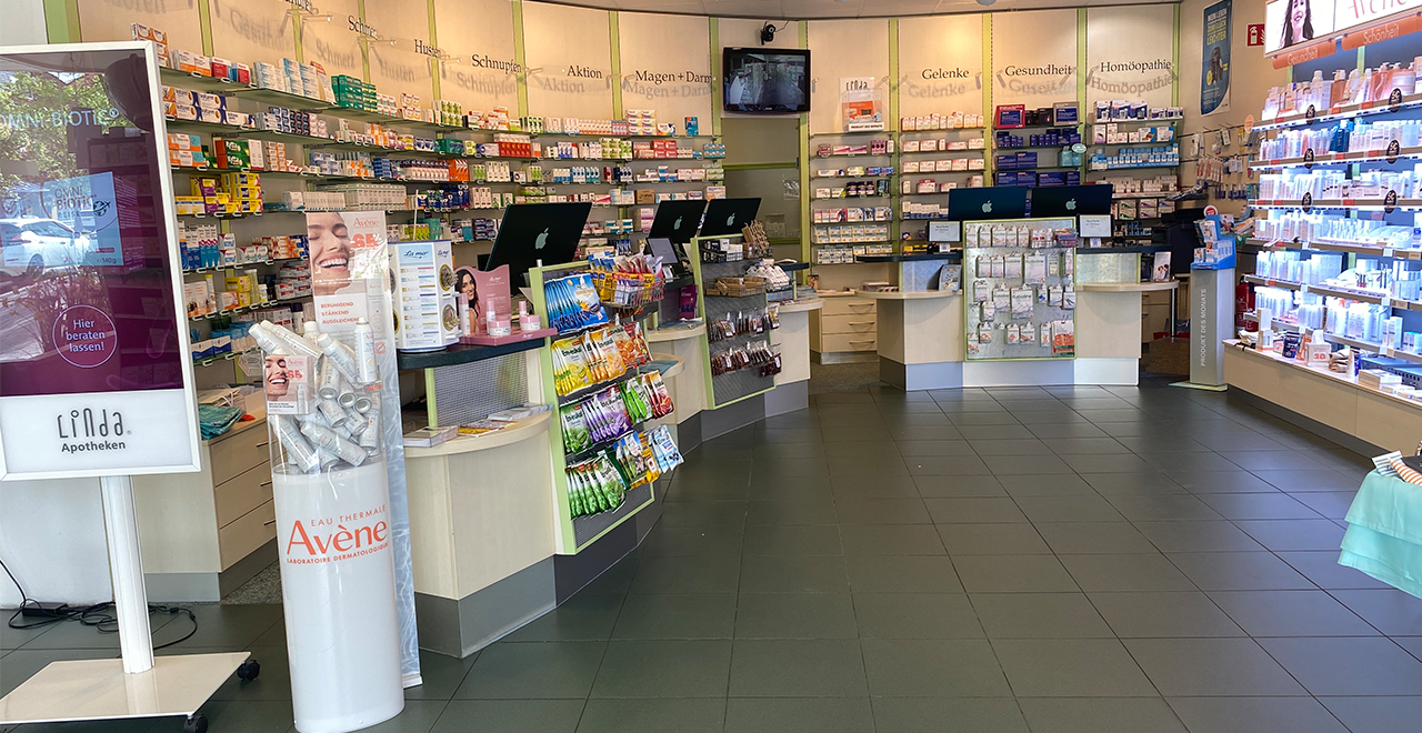 Heller, moderner Verkaufsraum einer Apotheke mit zahlreichen Regalen, gefüllt mit Medikamenten und Pflegeprodukten. In der Mitte stehen Verkaufstheken und Displays, auf der linken Seite ein digitaler Infoständer mit Werbung.