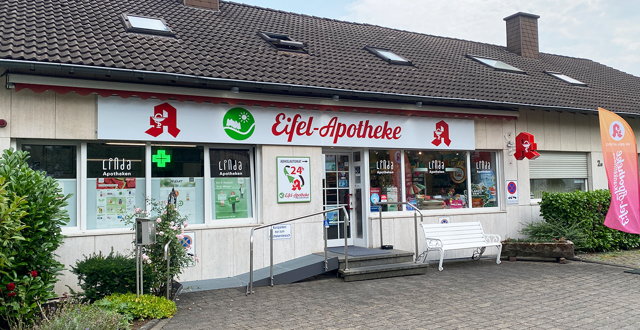 Außenansicht der Eifel-Apotheke mit barrierefreiem Zugang. Das Geschäft hat ein großes Schild mit dem Schriftzug „Eifel-Apotheke“ und einem roten Logo, das einen Mörser und Stößel zeigt. Ein grünes, leuchtendes Kreuz über dem Eingang kennzeichnet die Apotheke. Im Schaufenster sind Werbeanzeigen und das Logo von „Linda Apotheken“ zu sehen. Auf der rechten Seite befindet sich eine Bank vor dem Eingang und eine Rampe, die den barrierefreien Zugang ermöglicht. Ein 24-Stunden-Automat wird ebenfalls angezeigt.