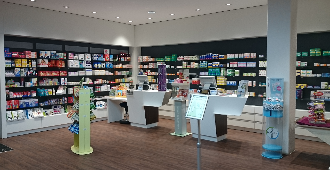 Innenraum einer Apotheke mit Regalen voller Medikamente, Produkten zur Körperpflege und einer modernen Theke. Kunden und Apotheker können im Hintergrund gesehen werden.