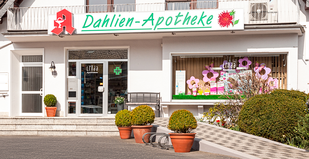Außenansicht der Dahlien-Apotheke mit dem grünen Schriftzug über dem Eingang. Die Apotheke befindet sich in einem hell verputzten Gebäude mit Balkon. Links und rechts vom Eingang stehen große Blumentöpfe mit Buchsbäumen. Das Schaufenster ist mit pinken Blumen und Produktdekoration zum Thema Frühling oder Allergiezeit gestaltet. Eine Bank steht vor dem Eingang.
