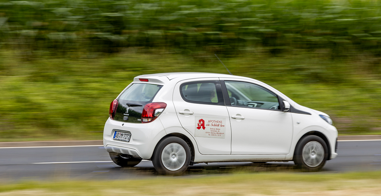 Weißes Peugeot-Auto mit Apotheken-Logo und -Adresse auf der Tür fährt auf einer Straße. Der Hintergrund ist aufgrund der Bewegung unscharf.