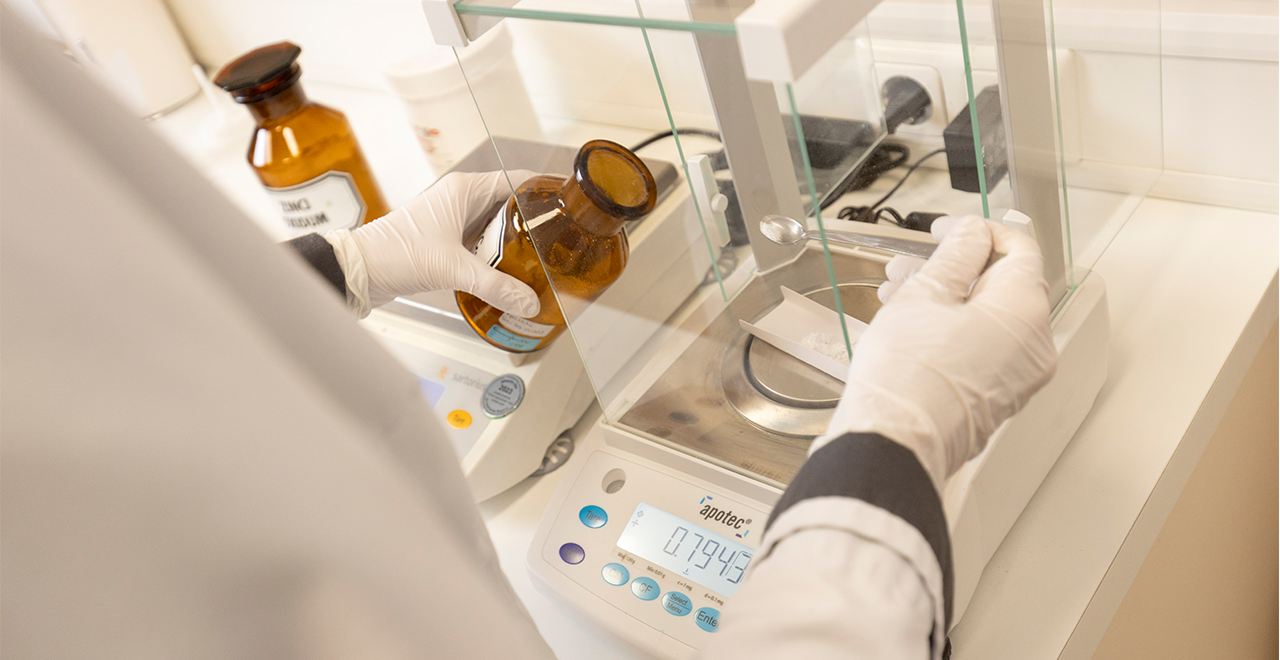 Eine Person in weißem Laborkittel und Handschuhen wiegt eine pulverförmige Substanz mit einer hochpräzisen Laborwaage. Daneben steht eine braune Apothekerflasche mit Beschriftung.