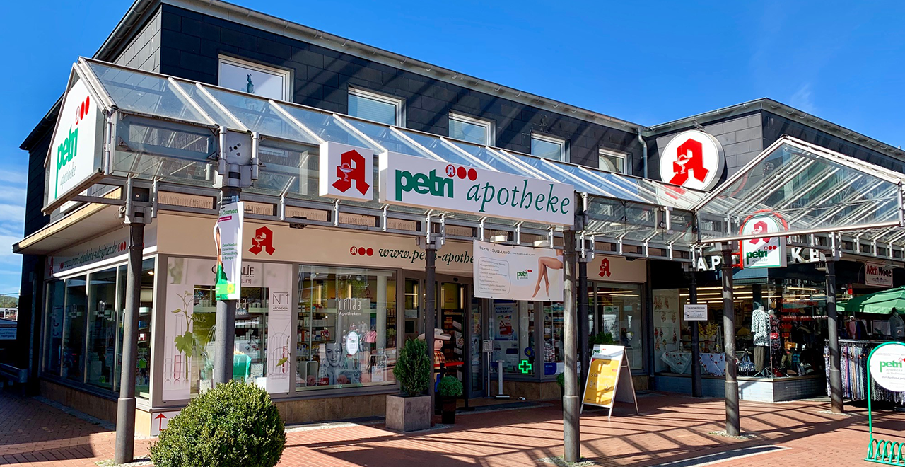 Foto einer Apotheke mit dem Namen ‚petri apotheke‘, das Gebäude hat ein modernes Design mit großen Fenstern und einem überdachten Eingangsbereich. Das Apotheken-Logo, ein rotes A mit einer Schale und Schlange, ist mehrfach sichtbar. Vor der Apotheke steht ein kleiner, runder grüner Busch. Der Himmel ist klar und blau.