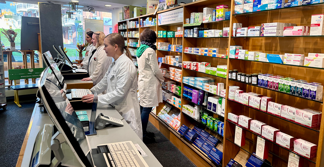 Mehrere Mitarbeitende in weißen Kitteln stehen hinter einer Verkaufstheke in einer Apotheke. Sie bedienen Kund*innen an Computerkassen. Im Hintergrund befinden sich gut sortierte Regale mit Medikamentenschachteln und Apothekenprodukten. Durch die großen Fenster fällt Tageslicht in den Raum.