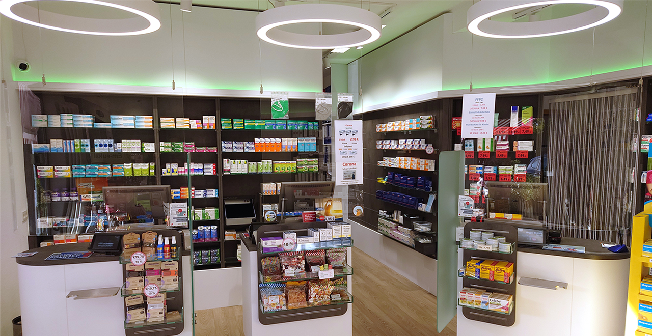Innenansicht einer modernen Apotheke mit dunklen Regalen und grüner LED-Beleuchtung. In der Mitte ein weißer Verkaufstresen mit einem Computer und Verkaufsständern. Rechts im Vordergrund ein Aufsteller, im Hintergrund zahlreiche Pflege- und Gesundheitsprodukte ordentlich einsortiert.