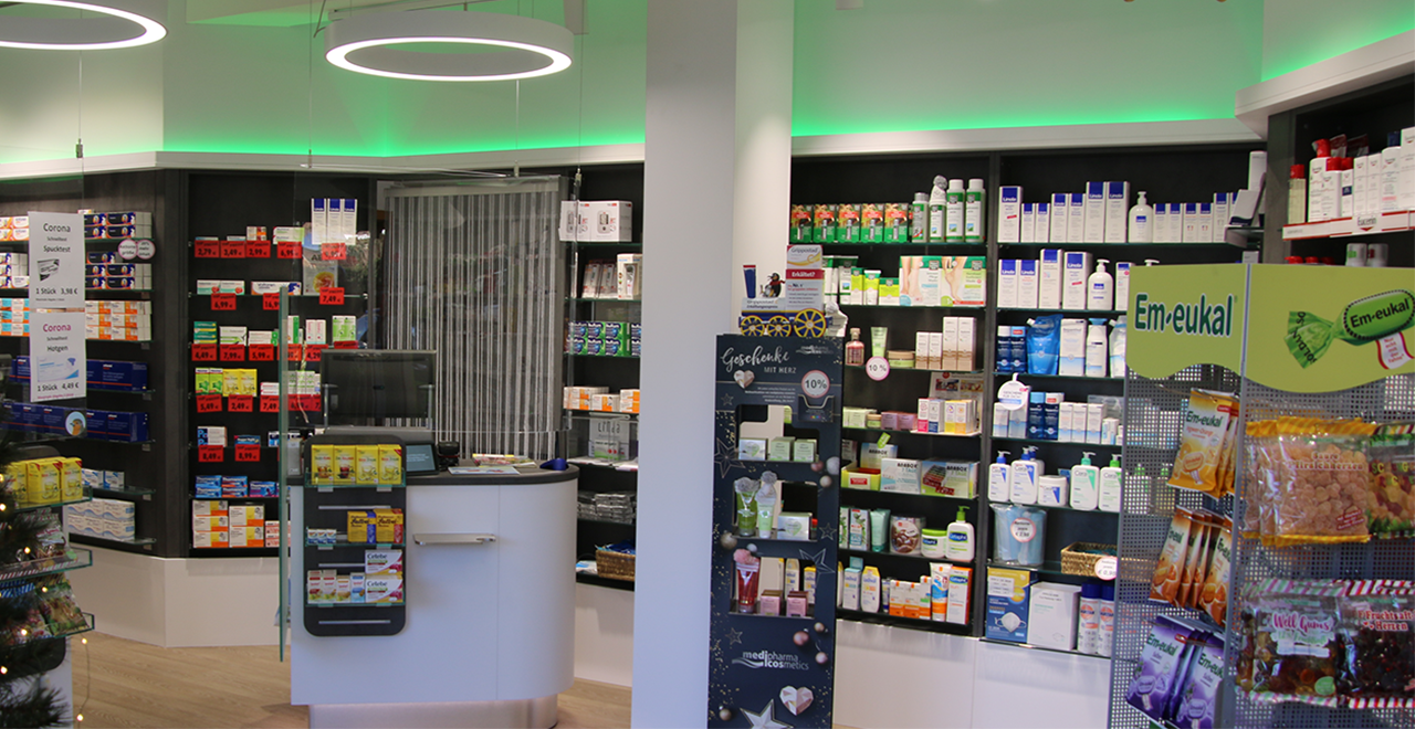Auf dem Bild sind zwei modernen Verkaufstresen, darüber kreisrunde Deckenleuchten. Die Regale im Hintergrund sind gefüllt mit Arzneimitteln und Gesundheitsprodukten. Auf den Tischen befinden sich kleine Produktständer und Aktionsangebote. Die Umgebung wirkt hell, aufgeräumt und einladend.