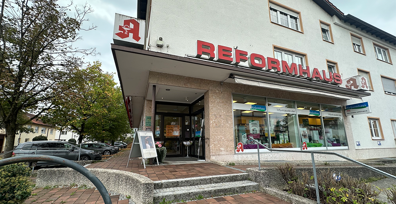 Eingang zu einem Reformhaus und einer Apotheke in einem Eckgebäude mit Glasfassade, Rampe und Blumen vor dem Eingang.