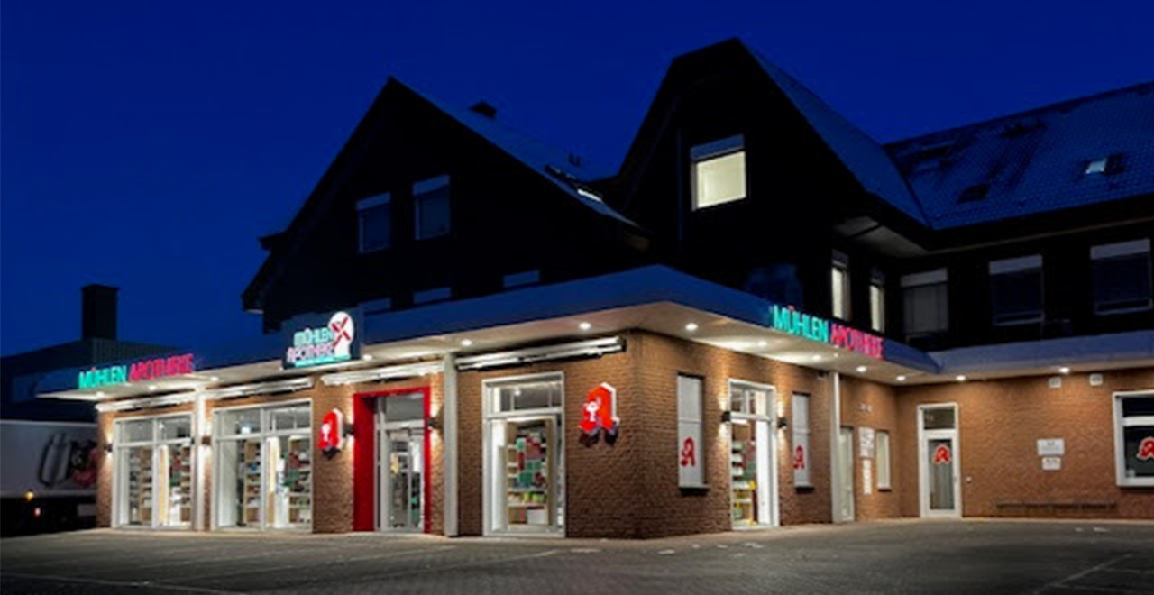 Außenansicht der Mühlen-Apotheke bei Dunkelheit.
Die beleuchteten Schriftzüge, Schaufenster und der überdachte Eingangsbereich heben sich vom dunklen Backsteingebäude ab.
Vor der Apotheke befindet sich ein Parkplatz.