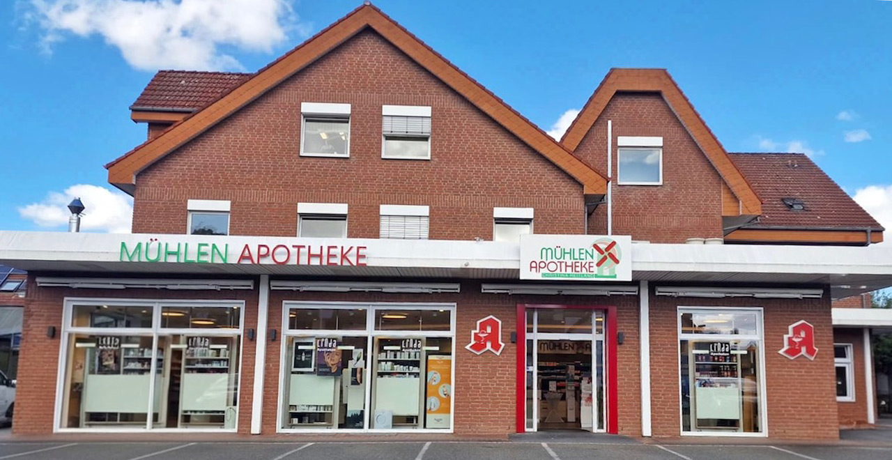 Außenansicht der Mühlen-Apotheke bei Tageslicht.
Das Backsteingebäude verfügt über große Schaufenster, einen zentralen barrierefreien Eingang und gut sichtbare grüne und rote Schriftzüge mit dem Namen der Apotheke.
Vor dem Gebäude befindet sich ein Parkplatz.