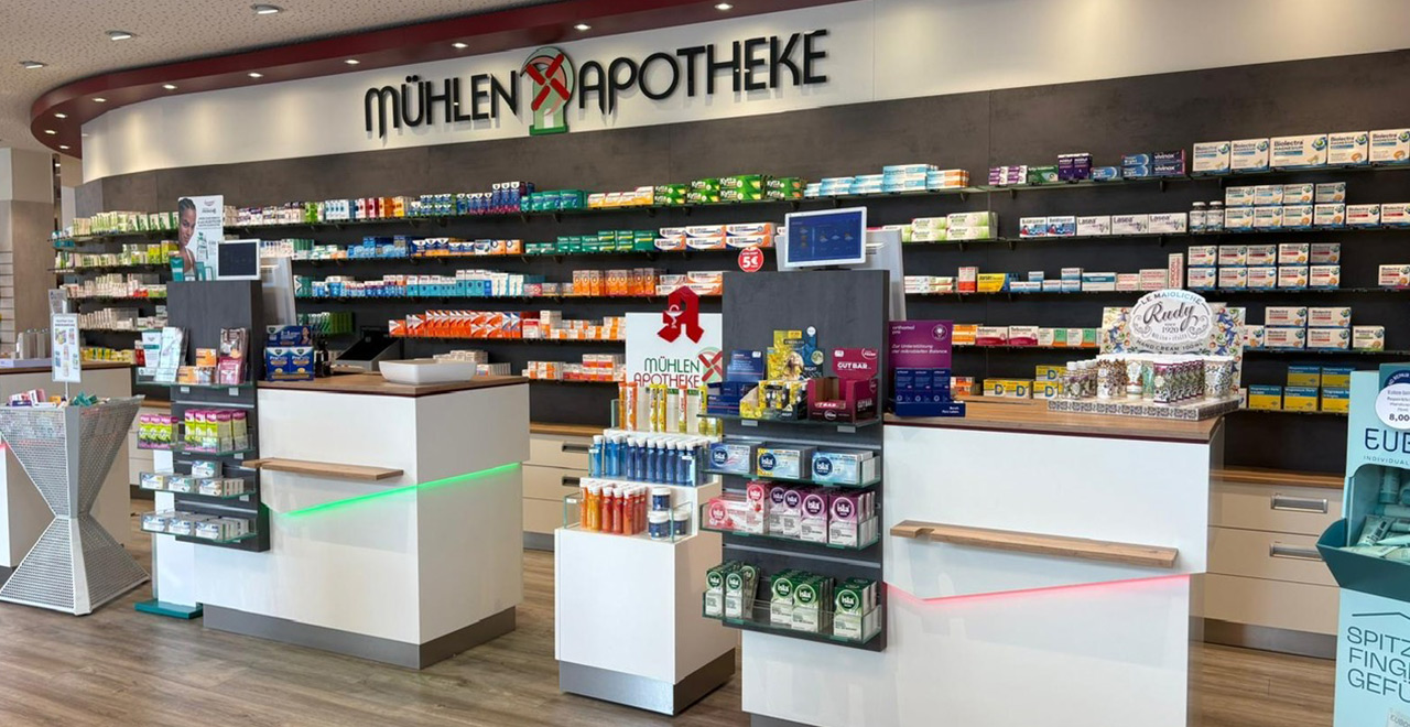 Innenansicht der Mühlen-Apotheke mit weißen Verkaufstresen, beleuchteten Regalen und einer großen Auswahl an Arzneimitteln, Gesundheitsprodukten und Pflegeartikeln.
Über den Regalen ist das Logo der Mühlen-Apotheke angebracht.