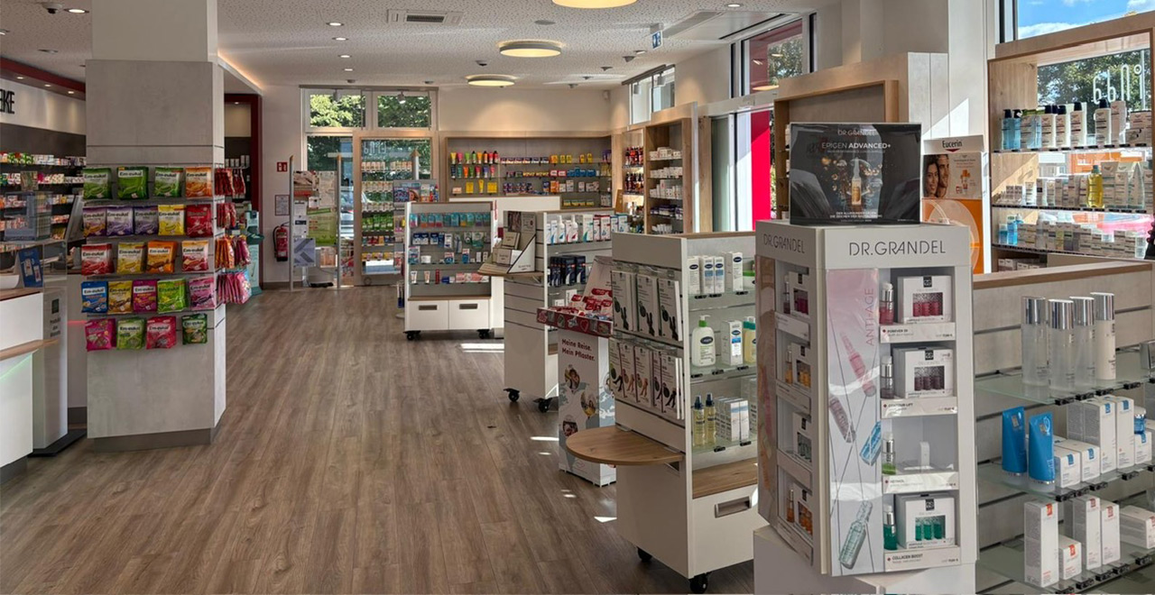 Innenansicht des Verkaufsraums der Mühlen-Apotheke mit breiten Holzoptik-Laufwegen, Regalen mit Arzneimitteln und Pflegeprodukten sowie Präsentationsständern für Kosmetik und Hautpflege.
Große Fenster sorgen für Tageslicht im hell gestalteten Raum.