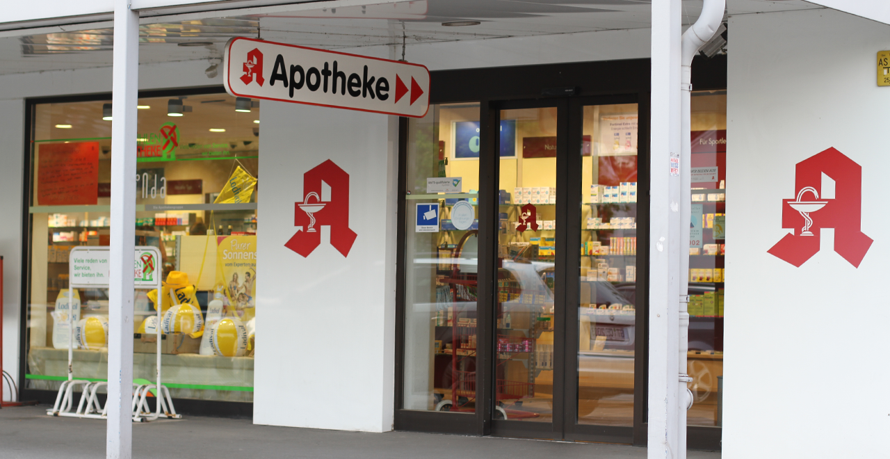 Außenansicht einer Apotheke mit Eingangstür, großer Beschilderung ‚Apotheke‘ und rotem Apotheken-Symbol. Durch die Glasfront sind Regale und Produkte im Verkaufsraum sichtbar.
