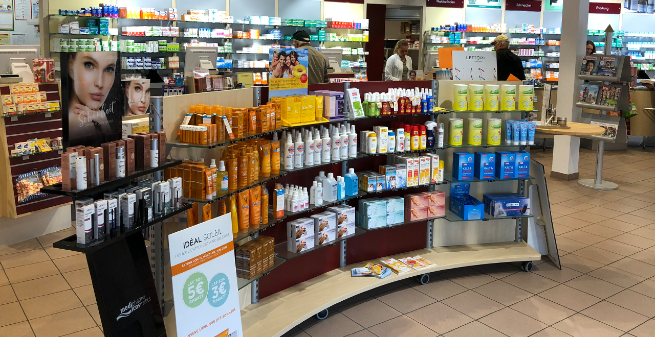 Innenansicht einer Apotheke mit Verkaufsregalen voller Pflegeprodukte, Sonnenschutz, Körperpflege und weiterer Apothekenartikel. Im Hintergrund befinden sich Beratungstheken und weitere Regale.