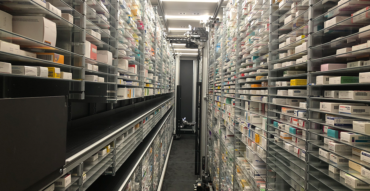 Automatisiertes Medikamentenlager einer Apotheke mit hohen, dicht gefüllten Regalen und einem Transportsystem in der Mitte.