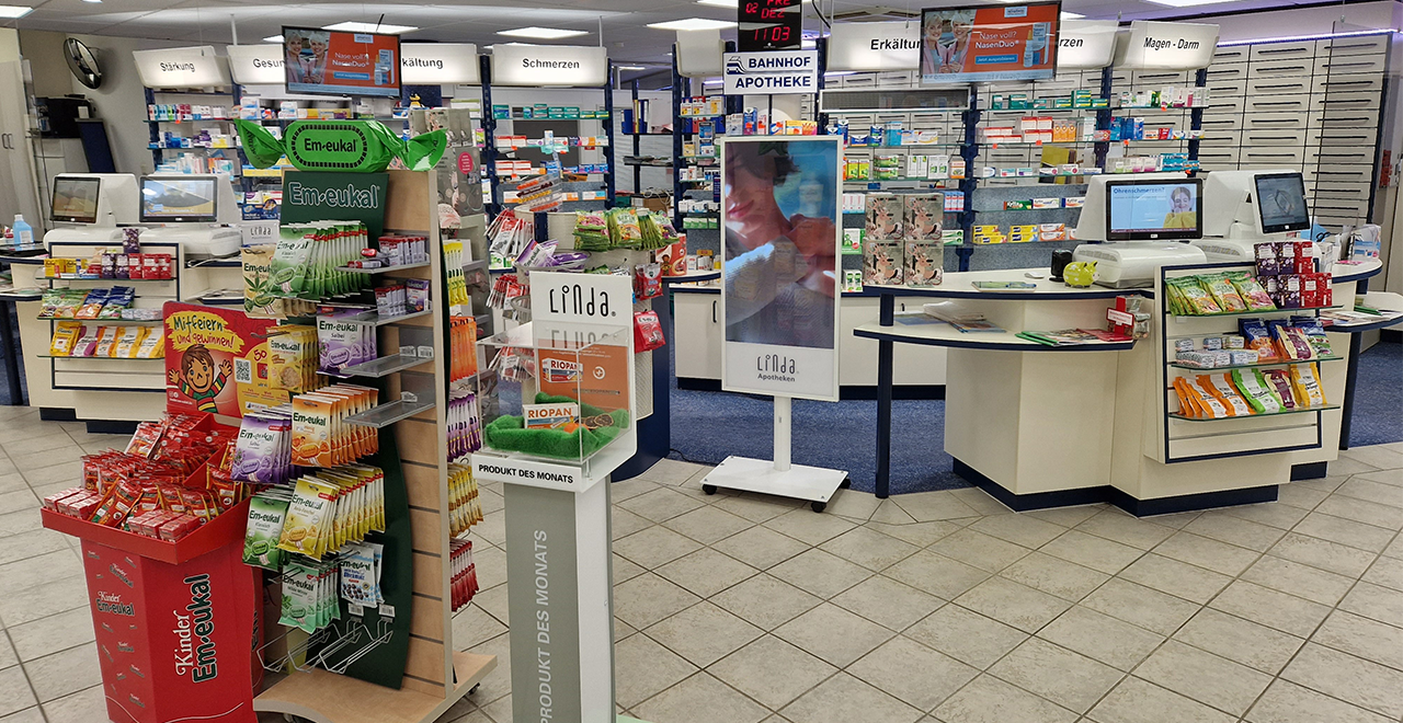Die Displays mit Em-eukal Produkten und das „Produkt des Monats“ stehen im Vordergrund. Im Hintergrund befinden sich die Kassen und Regale mit Medikamenten. Zwei große Monitore an der Decke zeigen Werbeinhalte und die Uhrzeit.