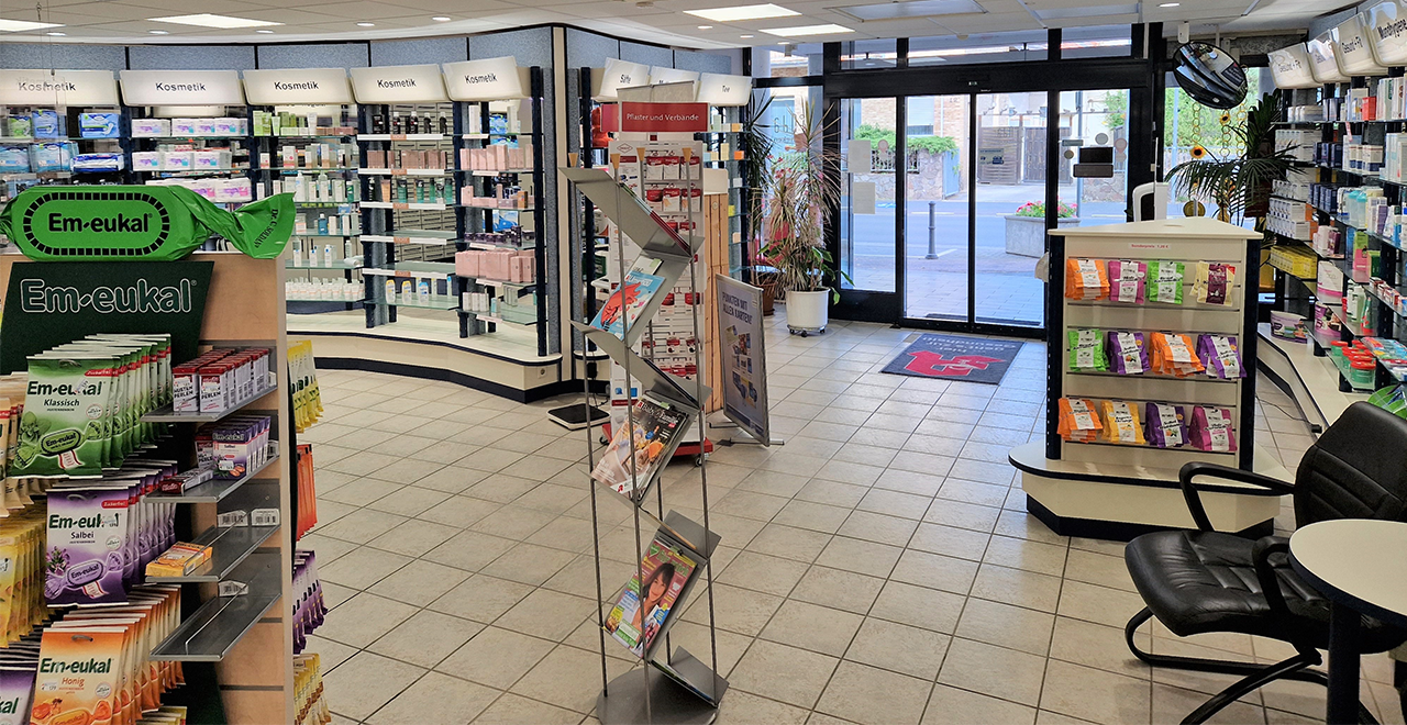 Blick aus dem hinteren Bereich der Apotheke Richtung Eingang. Links Regale mit Em-eukal Bonbons und Kosmetikprodukten. Rechts eine Sitzgelegenheit mit Tisch. In der Mitte ein Prospektständer mit verschiedenen Broschüren. Im Hintergrund sind die automatischen Eingangstüren zu sehen.