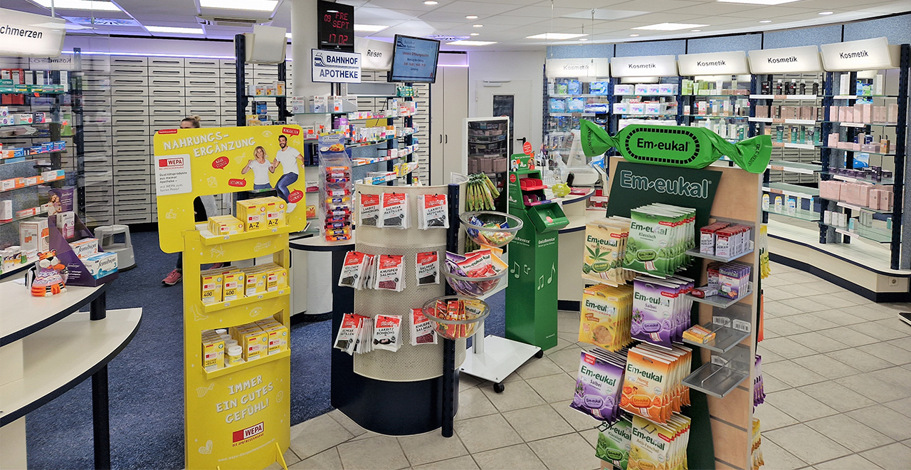 Innenansicht der Apotheke mit Fokus auf Verkaufsständer für Nahrungsergänzungsmittel, Lakritzprodukte und Em-eukal Bonbons. Im Hintergrund befinden sich Medikamentenregale, eine Monitoranzeige mit Uhrzeit sowie ein Bereich mit Kosmetikprodukten an der Wand.