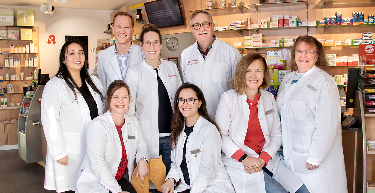 Acht Apothekenmitarbeitende – sechs Frauen und zwei Männer – stehen und sitzen lächelnd in einer hellen Apotheke. Sie tragen weiße Kittel und blicken in die Kamera. Hinter ihnen sind Regale mit verschiedenen Medikamenten und Gesundheitsprodukten zu sehen. Die Atmosphäre wirkt freundlich und professionell.