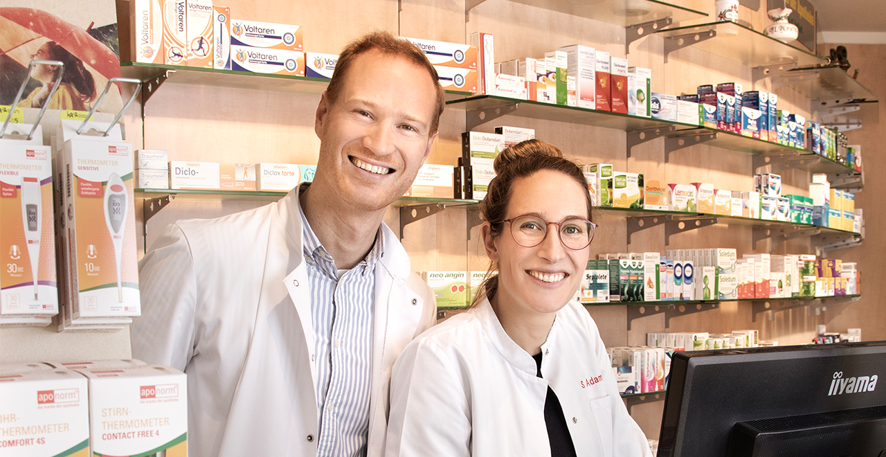 Zwei Apothekenmitarbeitende – eine Frau und ein Mann – stehen nebeneinander hinter einem Verkaufstresen und lächeln in die Kamera. Beide tragen weiße Kittel. Im Hintergrund befinden sich gut gefüllte Regale mit Medikamenten und Gesundheitsartikeln.