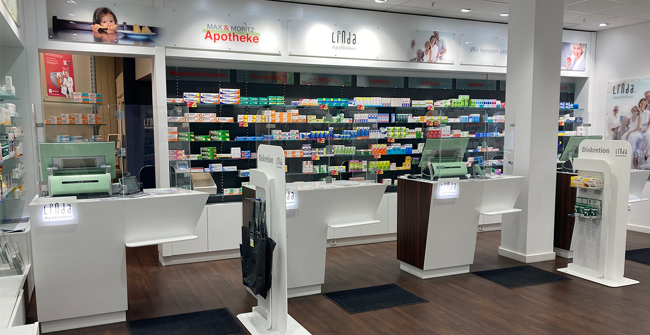 Ein moderner, heller Innenraum einer Apotheke mit weißen Verkaufstresen und transparenten Schutzscheiben. Hinter den Tresen befinden sich hohe Regale, gefüllt mit farblich sortierten Medikamentenschachteln. Über den Regalen hängen Leuchtkästen mit Fotos von Menschen und der Aufschrift „Max & Moritz Apotheke“ sowie „Linda Apotheken“. Der Boden ist dunkel, und einzelne kleine Warenträger stehen im Raum. Die Atmosphäre wirkt ordentlich, freundlich und barrierefrei gestaltet.