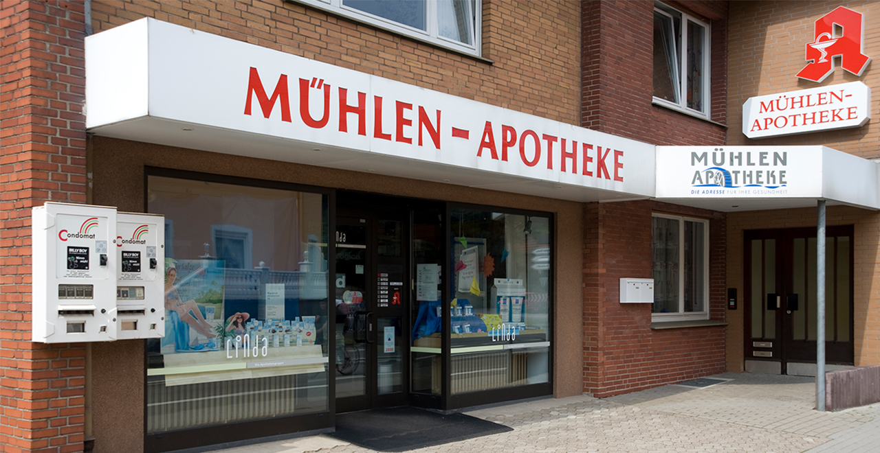 Außenansicht der Mühlen-Apotheke in einem Backsteingebäude. Über dem Eingang prangt ein weißes Apothekenschild mit roter Schrift „MÜHLEN-APOTHEKE“ und dem roten Apotheken-A auf der rechten Seite. Rechts neben dem Haupteingang ein weiteres Schild mit modernem Logo und dem Slogan „Die Adresse für Ihre Gesundheit“. Im Schaufenster sind verschiedene Produkte und Werbematerialien ausgestellt.